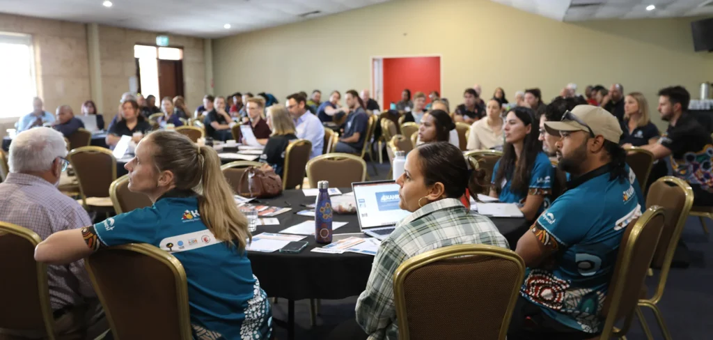 Diverse attendees at a Kimberley Aboriginal Health Research Alliance (KAHRA) workshop, many wearing teal shirts with Indigenous art, seated at round tables in a conference room.